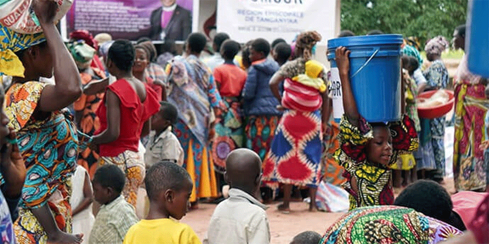 A young girl carries a bucket of supplies on her head amid a crowd of beneficiaries gathered in front of banners from UMCOR and the Tanganyika Episcopal Area. The distribution of relief kits aims to restore the dignity of families in Nyunzu, who are struggling to survive in the face of food insecurity and disease resulting from recent floods and conflicts in eastern DRC. Photo courtesy of the Disaster Management Office of the Tanganyika Episcopal Area.