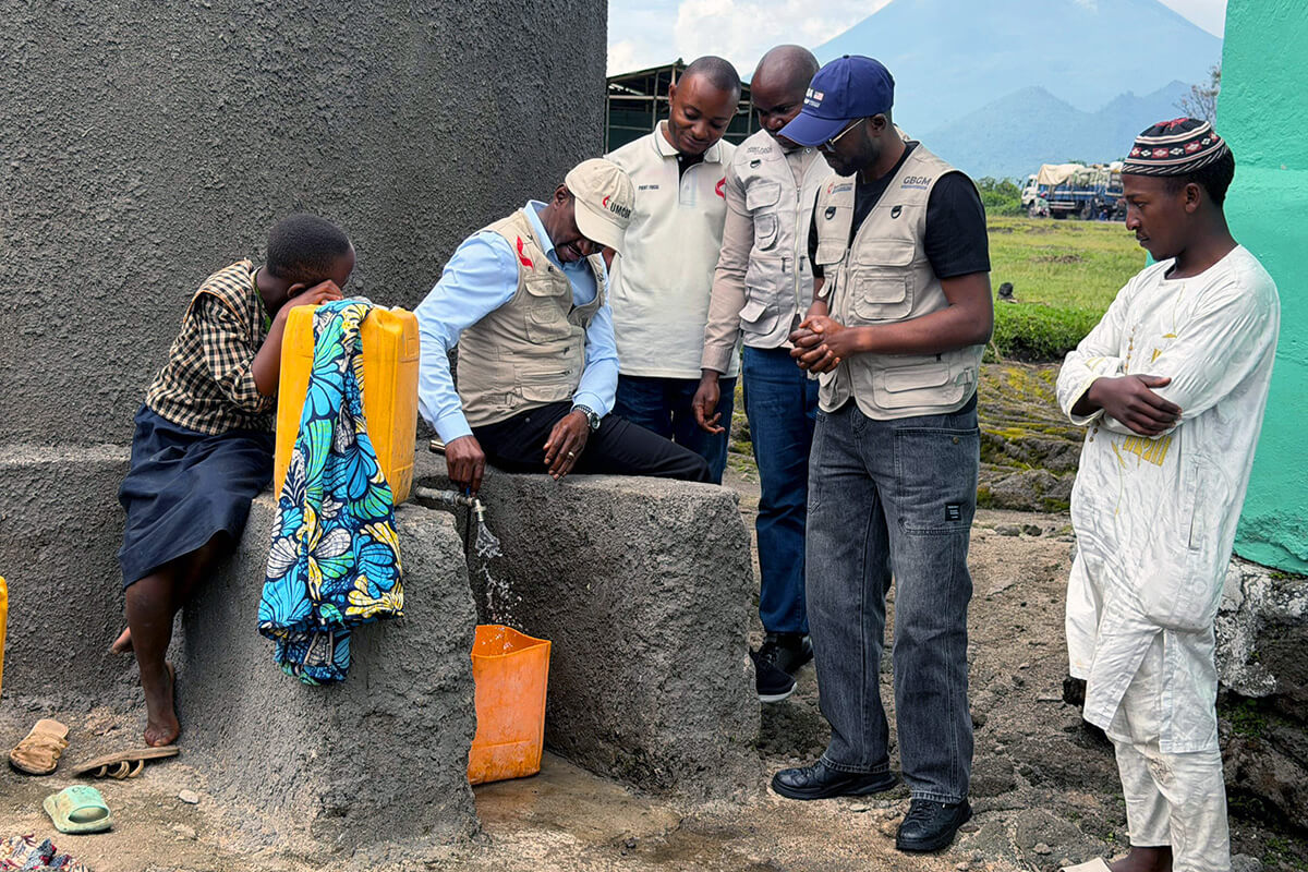 Jean Tshomba, Coordinateur du Bureau de gestion des catastrophes du Comité Méthodiste Uni pour les Secours (UMCOR) dans l’Est du Congo, ouvre le robinet d’une nouvelle citerne d’eau de pluie à Kibumba, au Congo, sous le regard du Dr Damas Lushima (coiffé d’un chapeau bleu), des membres de l’équipe et d’une jeune fille du village. Grâce à un financement de UMCOR, 24 citernes de collecte d’eau de pluie ont été installées afin d’assurer un accès vital à l’eau potable dans ce village isolé, dépourvu de sources d’eau de surface et fragilisé par les récents déplacements de population. Photo de Jolie Shabani, UM News.
